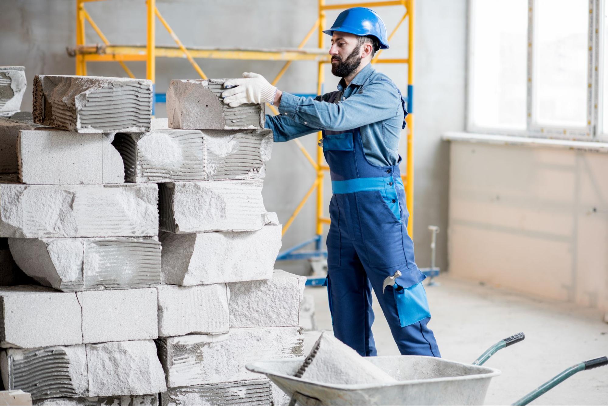 A construction worker in safety gear stacks heavy stone blocks during an indoor building renovation project.