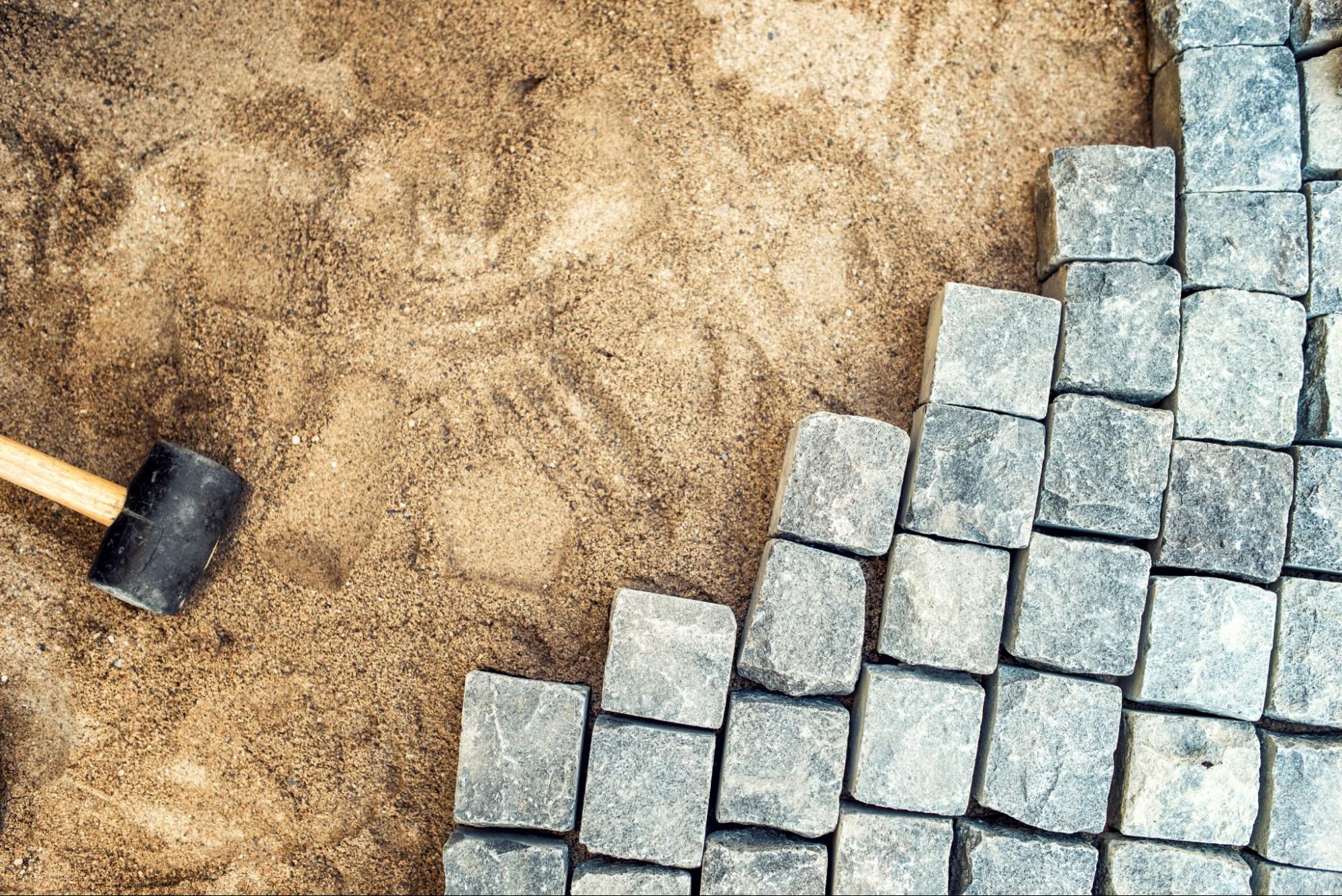 Top view of paving tools and cobble stones arranged on sand during a pavement installation.